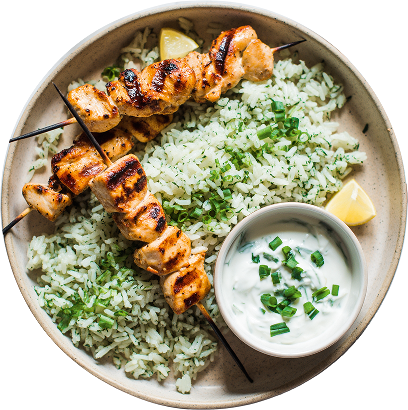 Zitronen-Hähnchenspieße mit Kräuterreis und cremigem Joghurt-Dip in einer Schale, garniert mit frischen Kräutern und Zitronenspalte.