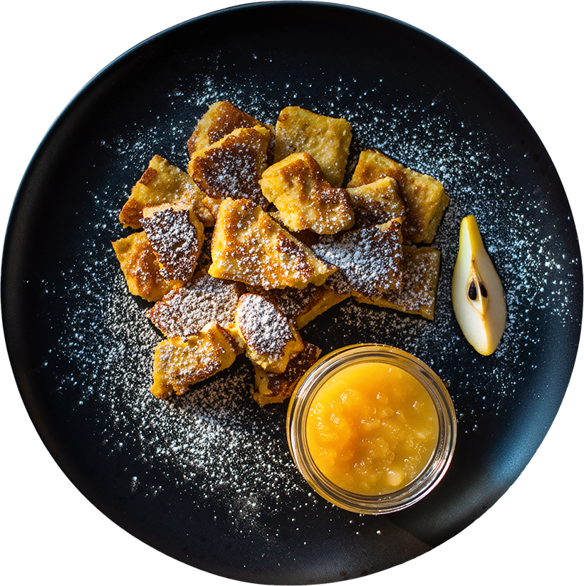 Kaiserschmarrn mit Puderzucker und warmem Birnenkompott auf einem schwarzen Teller angerichtet.
